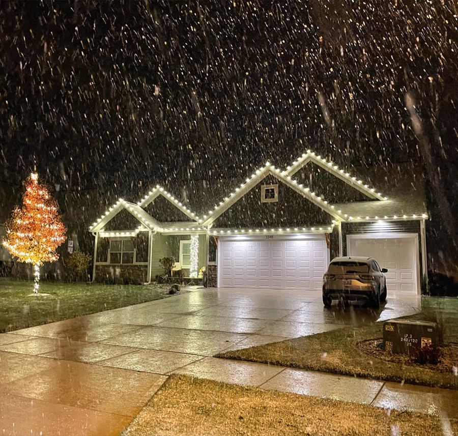 Single-story home with roofline lights and a lit tree in falling snow